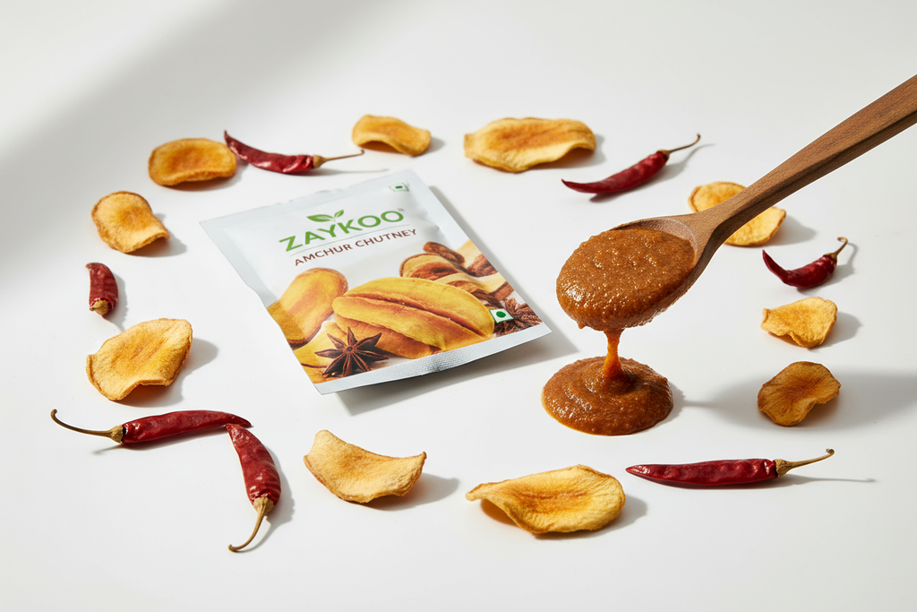 Zaykoo Amchur Chutney packet centered with a dripping spoon of chutney beside it. Dried mango slices and chillies scattered artistically.